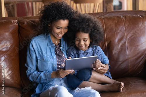 Smiling young African American mother and teen ethnic daughter relax on sofa at home use modern tablet gadget together. Happy ethnic mom and small girl child have fun rest with pad device.