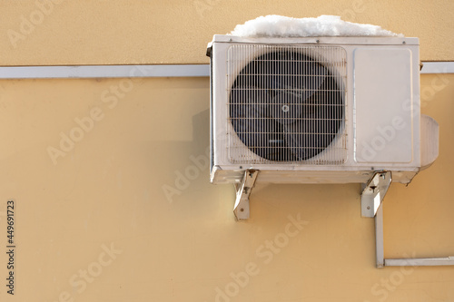 An outdoor air conditioner unit installed on the outer wall of a residential building. The fan and the radiator grille of the air conditioner are close-up.