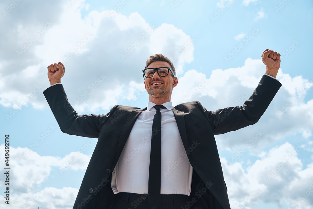 Happy mature man in full suit keeping arms raised while standing in front of blue sky outdoors Happy mature man in full suit keeping arms raised while standing in front of blue sky outdoors