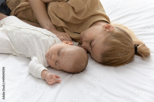 Young Caucasian mother and little baby infant kid child lying sleeping together in cozy white bed at home. Loving mom and small newborn daughter or son take nap fall asleep. Parenthood concept.