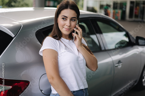 Fényképezés Attractive young woman using her phone and leaned on her car at gas station