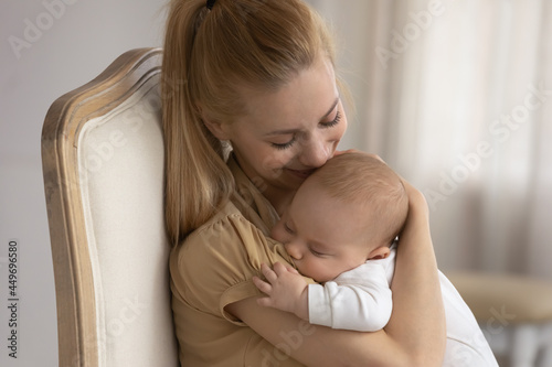 Happy young Caucasian mother hug caress little baby infant son or daughter sleeping in hands. Smiling mom sit in chair rest cuddle lull small newborn kid child relax in arms. Motherhood concept.