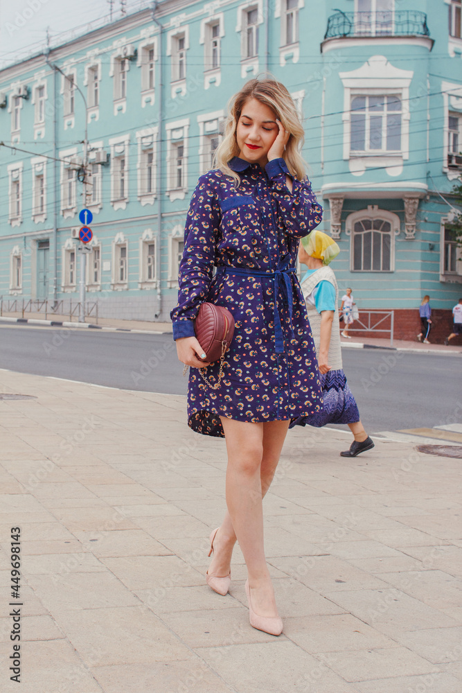 Young beautiful woman in blue dress with a round bag walking on the spring street. Urban background. Stylish Tourist girl enjoying walking the city during weekend trip.
