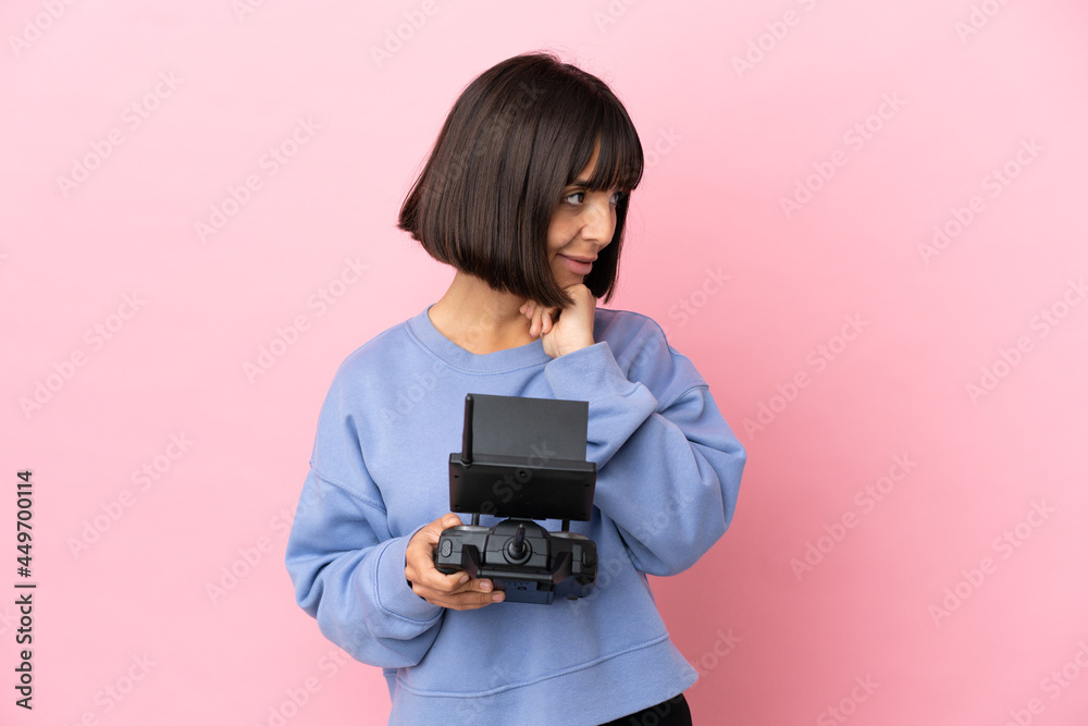 Young mixed race woman holding a drone remote control isolated on pink background looking up while smiling Young mixed race woman holding a drone remote control isolated on pink background looking up while smiling