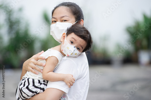 Asian mother wearing white face mask holding her little toddler baby girl up close who is her daughter wear baby face mask, look at camera, COVID-19 concept