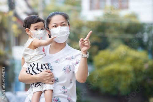 Asian mother wearing white face mask holding her little toddler baby girl who is her daughter wear baby face mask, pointing finger and looking out, COVID-19 concept