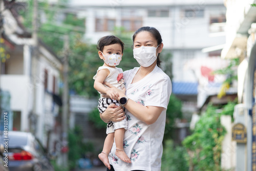 Asian mother wearing white face mask holding her little toddler baby girl who is her daughter wear baby face mask, look at camera, COVID-19 concept