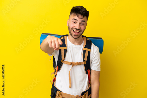 Young mountaineer caucasian man with a big backpack isolated on yellow background surprised and pointing front