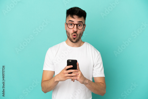 Young caucasian man isolated on blue background looking at the camera while using the mobile with surprised expression