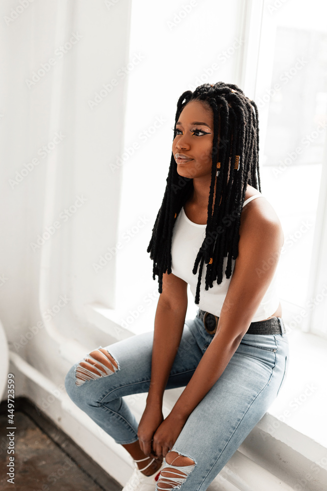 Gorgeous African American woman with cool dreadlocks in fashionable clothes  sits near window. Beautiful black afro girl with trendy hairstyle in  fashion summer outfit relaxes indoors. Stock Photo | Adobe Stock