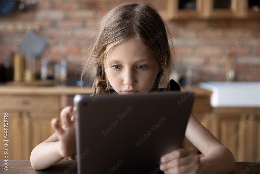 Focused gen Z little schoolkid using online app on tablet, virtual service on computer at home. Serious girl, child with digital device browsing internet, reading electronic book in kitchen Focused gen Z little schoolkid using online app on tablet, virtual service on computer at home. Serious girl, child with digital device browsing internet, reading electronic book in kitchen