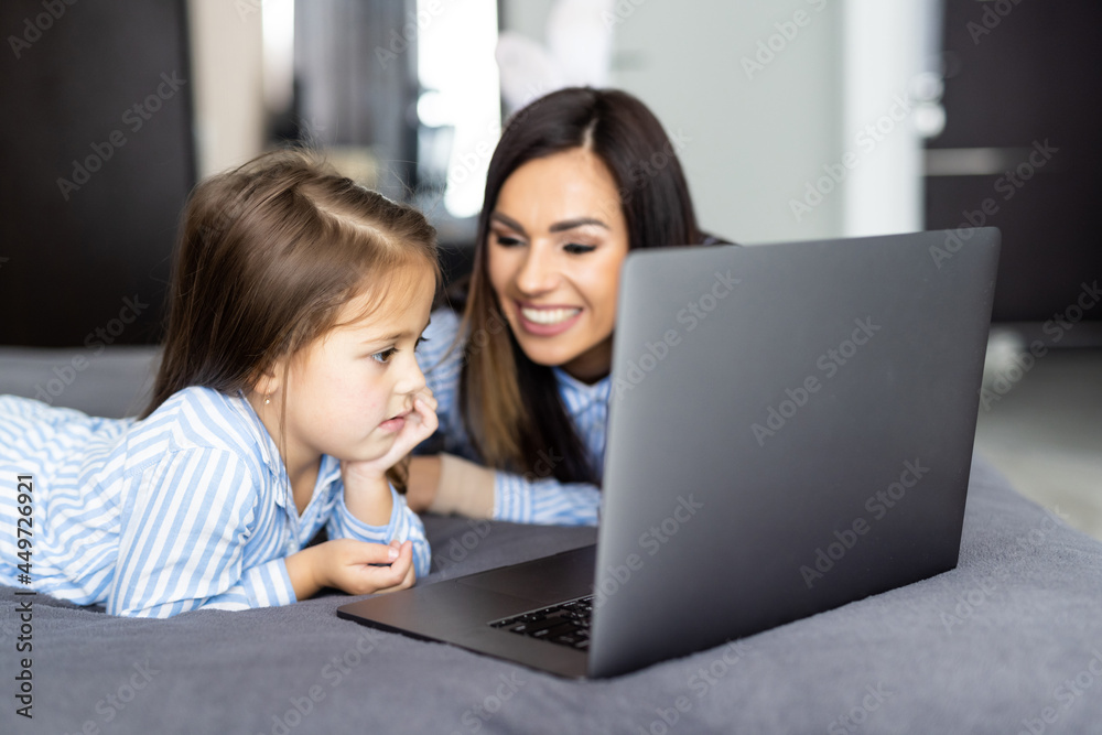 Portrait young happy smiling mother work from home with lovely daughter use laptop while together sitting on bed. Portrait young happy smiling mother work from home with lovely daughter use laptop while together sitting on bed.