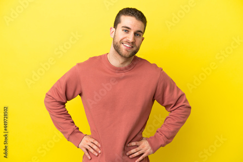Handsome blonde man over isolated yellow background posing with arms at hip and smiling
