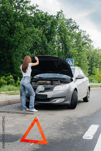 A young girl stands near a broken-down car in the middle of the highway and looks under the hood. Failure and breakdown of the car. Waiting for help. © Andrii