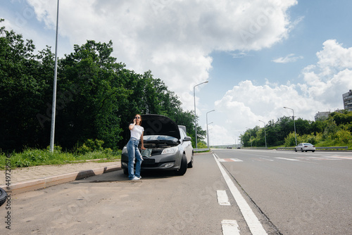 A young girl stands near a broken-down car in the middle of the highway and calls for help on the phone. Failure and breakdown of the car. Waiting for help.