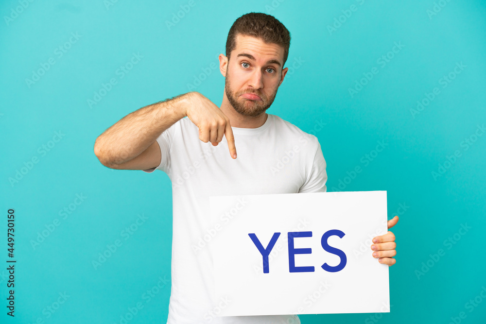 Handsome blonde man over isolated blue background holding a placard with text YES and pointing it Handsome blonde man over isolated blue background holding a placard with text YES and pointing it