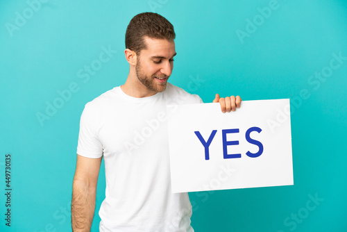 Handsome blonde man over isolated blue background holding a placard with text YES © luismolinero