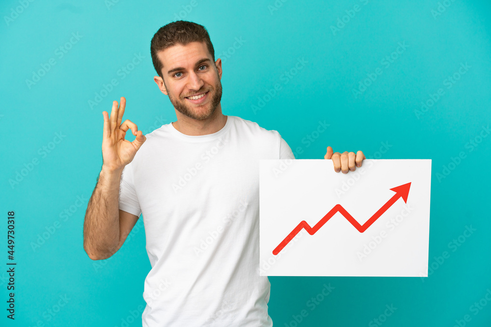 Handsome blonde man over isolated blue background holding a sign with a growing statistics arrow symbol with ok sign