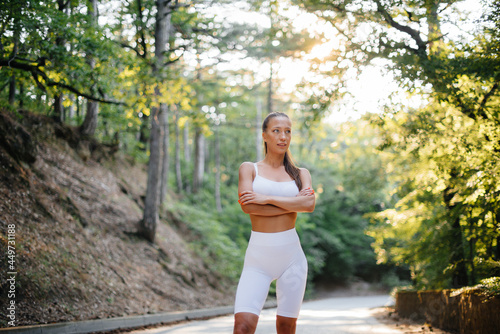 A young beautiful girl poses before running training, on the road in a dense forest, during sunset. A healthy lifestyle and running in the fresh air.