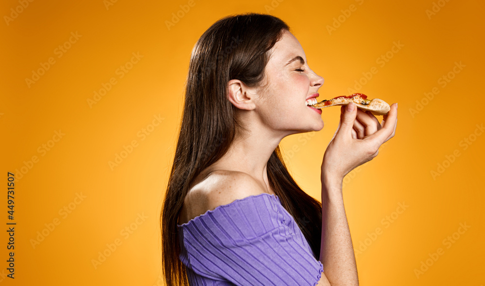 Profile portrait of young woman face biting slice of pizza, eating fastfood with satisfaction and smiling pleased, standing against orange background Profile portrait of young woman face biting slice of pizza, eating fastfood with satisfaction and smiling pleased, standing against orange background