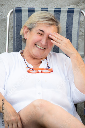 Blond woman sit laughing with her pink glasses on her neck over a grey background on a summer afternoon in Spain