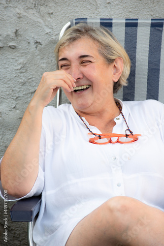 Blond woman sit laughing with her pink glasses on her neck over a grey background on a summer afternoon in Spain