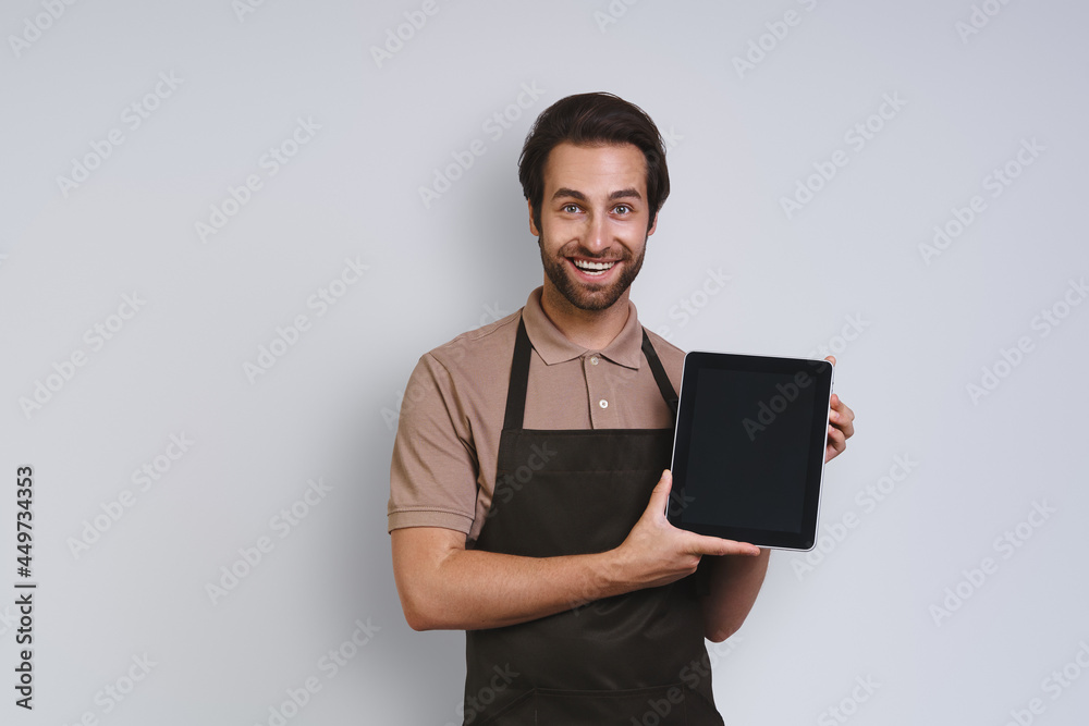 Cheerful young man in apron showing his digital tablet and smiling while standing against gray background