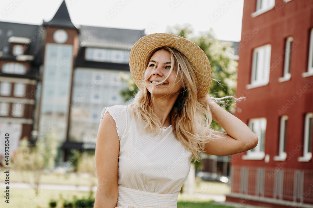A beautiful blonde walks through a European city. Woman in white dress and straw hat, she smiles and happy