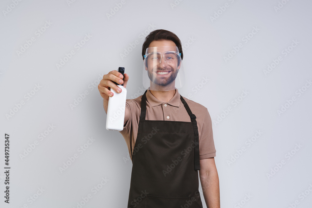 Handsome young man in apron wearing protective face shield and holding cleaning spray while standing against gray background Handsome young man in apron wearing protective face shield and holding cleaning spray while standing against gray background