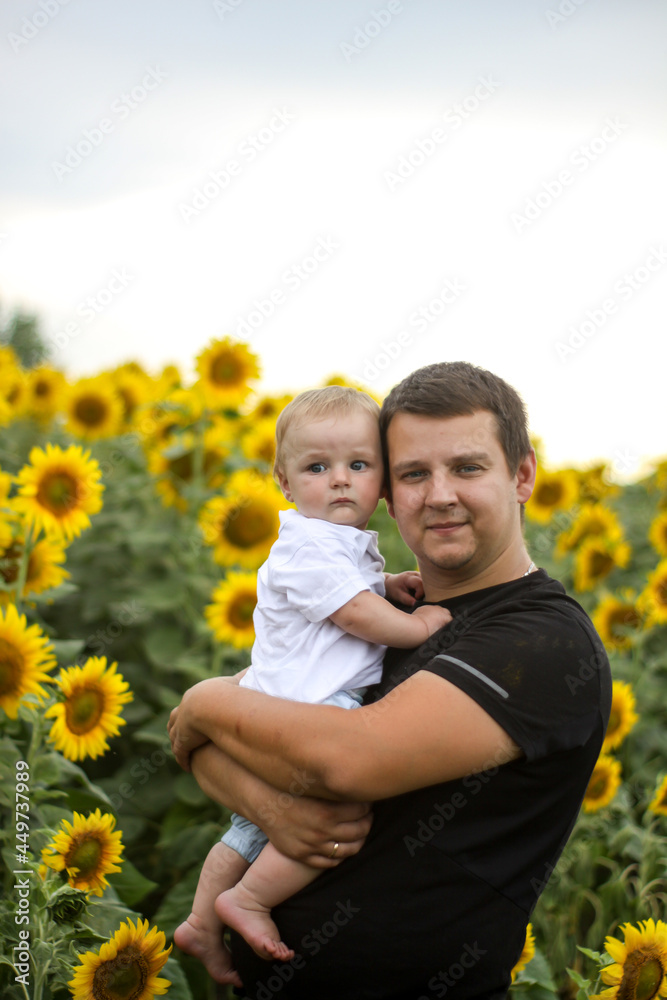 Fototapeta premium brunette male young dad with cute child blonde son on a field of sunflowers.