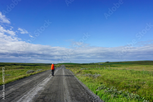 a tourist with an orange backpack on his back is walking along an empty dirt road, a hilly green area, a blue sky with clouds, a sunny day, the nature of Iceland