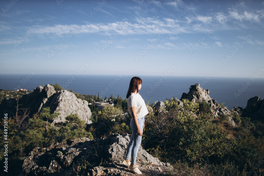 woman traveler in blue jeans and a white T-shirt with dark hair stands on a stone and looks at a rocky landscape by the black sea