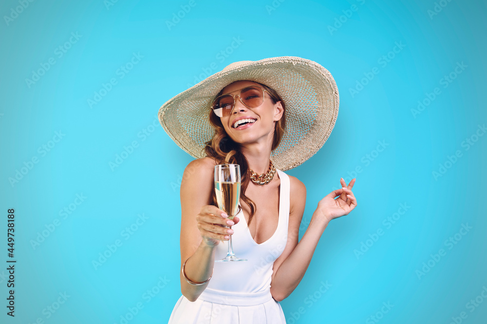 Fototapeta premium Beautiful young woman in elegant hat holding champagne flute and smiling while standing against blue background