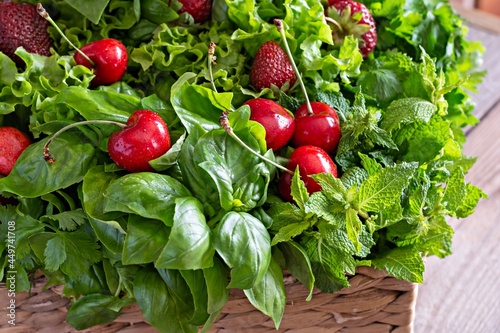 Fresh various fragrant herbs with cherries and strawberries in a wicker basket on a wooden background. Healthy food concept. Summer garden harvest.