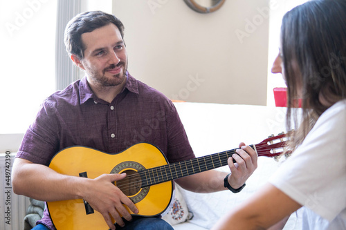 Young and attractive guy playing a romantic song in the guitar to his girlfriend while enjoying a day at home. Woman is surprised and happy listening to his song.