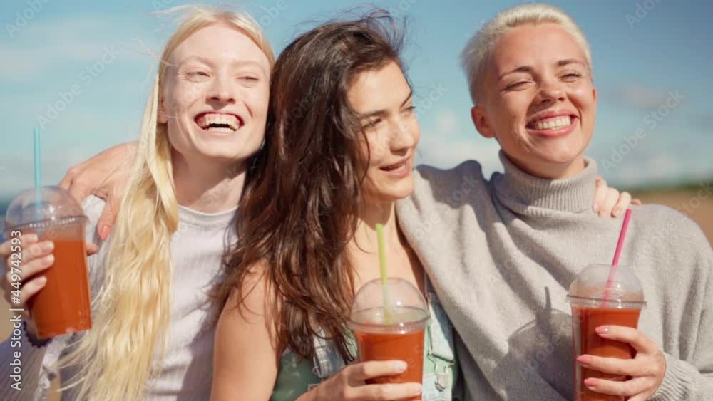 Slow motion waist up portrait of three happy young girlfriends or sisters hugging, talking and laughing while standing with smoothie on sea beach. Happy women enjoying vacation and friendship Slow motion waist up portrait of three happy young girlfriends or sisters hugging, talking and laughing while standing with smoothie on sea beach. Happy women enjoying vacation and friendship