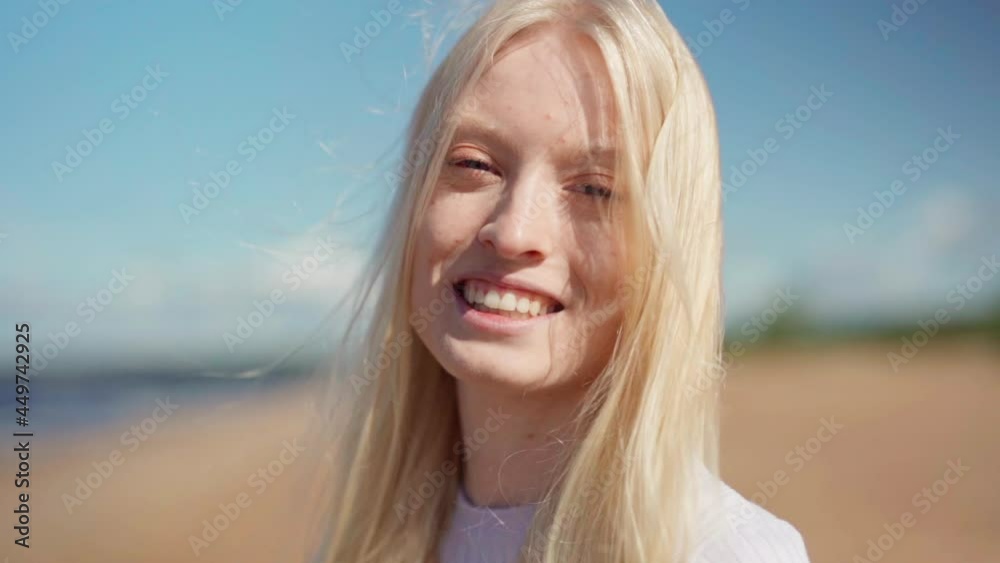 Closeup portrait face of happy beautiful young woman looking at camera smiling and laughing while posing on sea beach, long blonde hair waving in wind in slow motion. Travel and natural beauty concept Closeup portrait face of happy beautiful young woman looking at camera smiling and laughing while posing on sea beach, long blonde hair waving in wind in slow motion. Travel and natural beauty concept