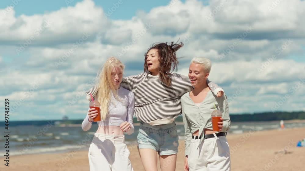 Slow motion shot of two laughing young women with cocktails walking along sea coast on beach. Happy girlfriend joining and hugging them, women raising arm and screaming for joy celebrating freedom Slow motion shot of two laughing young women with cocktails walking along sea coast on beach. Happy girlfriend joining and hugging them, women raising arm and screaming for joy celebrating freedom