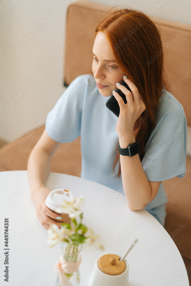 Vertical top view of happy young woman holding cup with hot coffee in hands talking on mobile phone sitting at desk in cafe. Pretty redhead Caucasian lady having leisure activity in coffee shop.