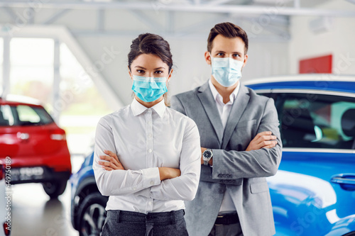 Two successful, proud car sellers standing with arms crossed in car salon and having face masks on faces.