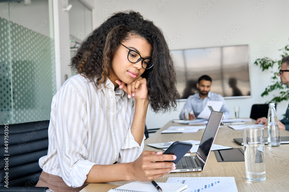 Young African American business woman using smartphone sitting in office at busy team meeting. Female manager employee working on phone and laptop technology corporate group briefing. Young African American business woman using smartphone sitting in office at busy team meeting. Female manager employee working on phone and laptop technology corporate group briefing.