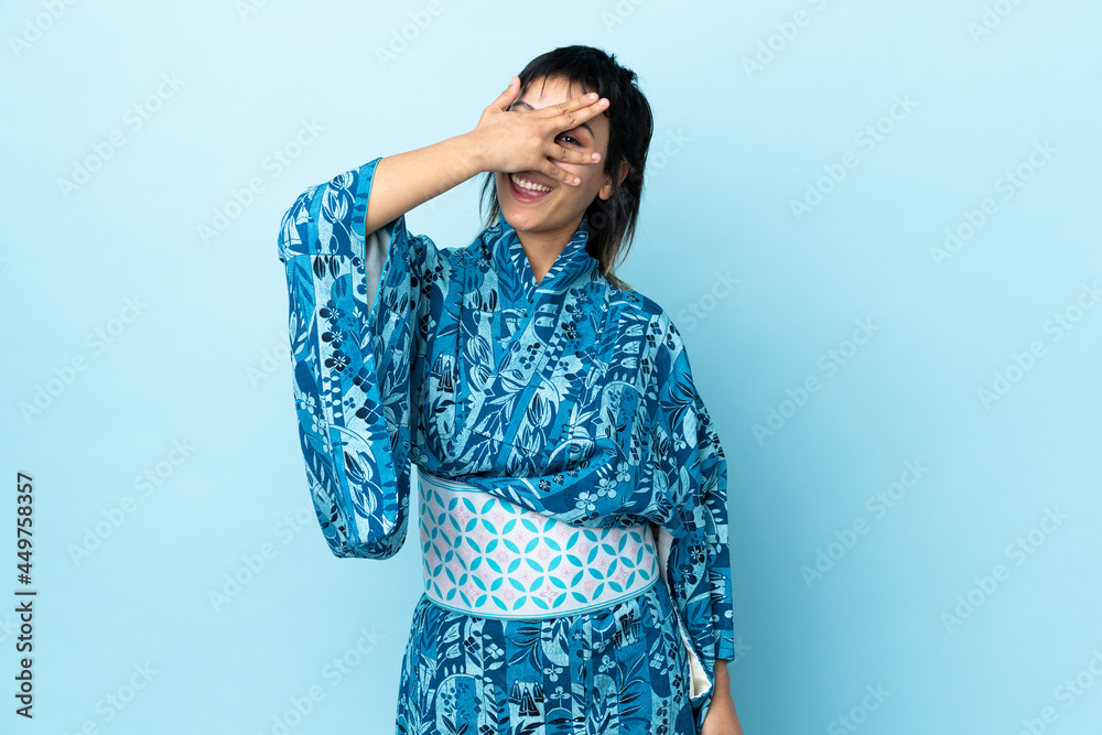 Young woman wearing kimono over isolated blue background covering eyes by hands and smiling Young woman wearing kimono over isolated blue background covering eyes by hands and smiling