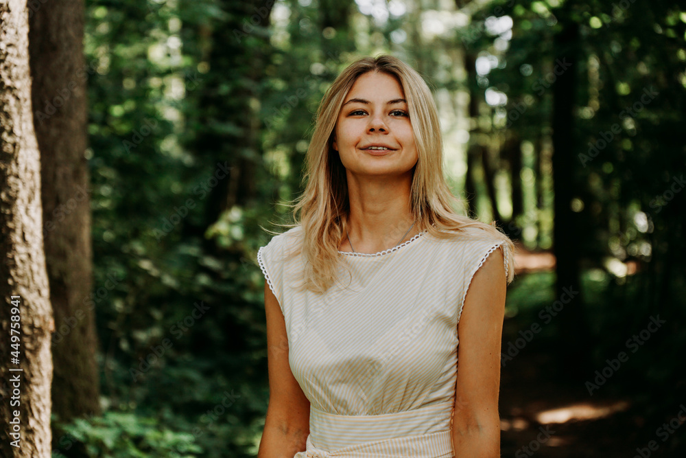 Attractive young woman enjoying her time outside in park. Young blonde in a white dress in a green park. Woman on the background of summer trees Attractive young woman enjoying her time outside in park. Young blonde in a white dress in a green park. Woman on the background of summer trees