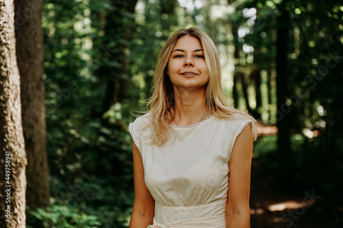 Attractive young woman enjoying her time outside in park. Young blonde in a white dress in a green park. Woman on the background of summer trees