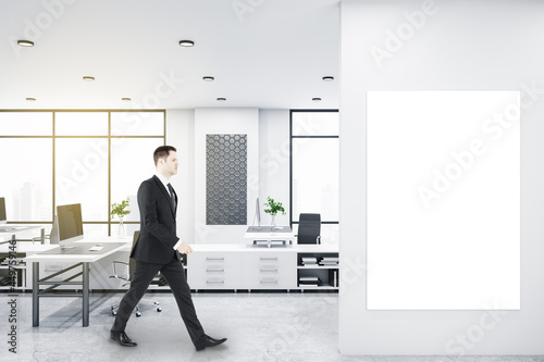 Businessman walking in bright coworking office interior with empty banner, window and city view, daylight, furniture and equipment. Mock up.