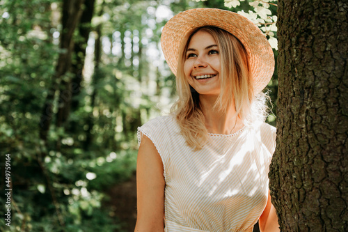 Beautiful girl in a straw hat and stylish clothes stands in a park near a tree. A woman in a white dress near a tree. Close portrait of happy girl
