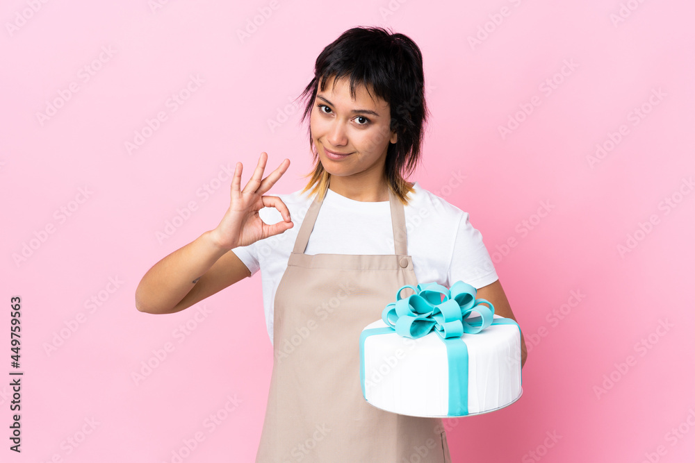 Uruguayan Pastry chef holding a big cake over isolated pink background showing an ok sign with fingers Uruguayan Pastry chef holding a big cake over isolated pink background showing an ok sign with fingers