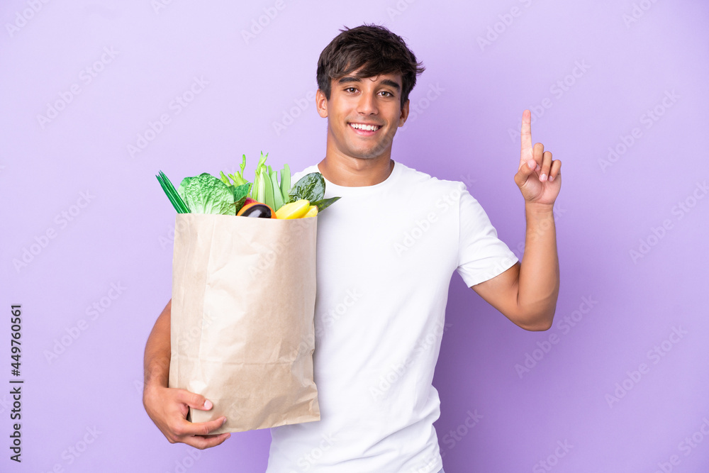Young man holding a grocery shopping bag isolated on purple background pointing up a great idea Young man holding a grocery shopping bag isolated on purple background pointing up a great idea