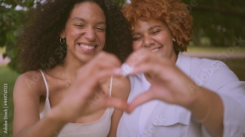 Young beautiful happy lesbian African American couple sitting on green grass showing love sign outside at nature summer park. LGBT community concept. Female friends smiling enjoying moments together.