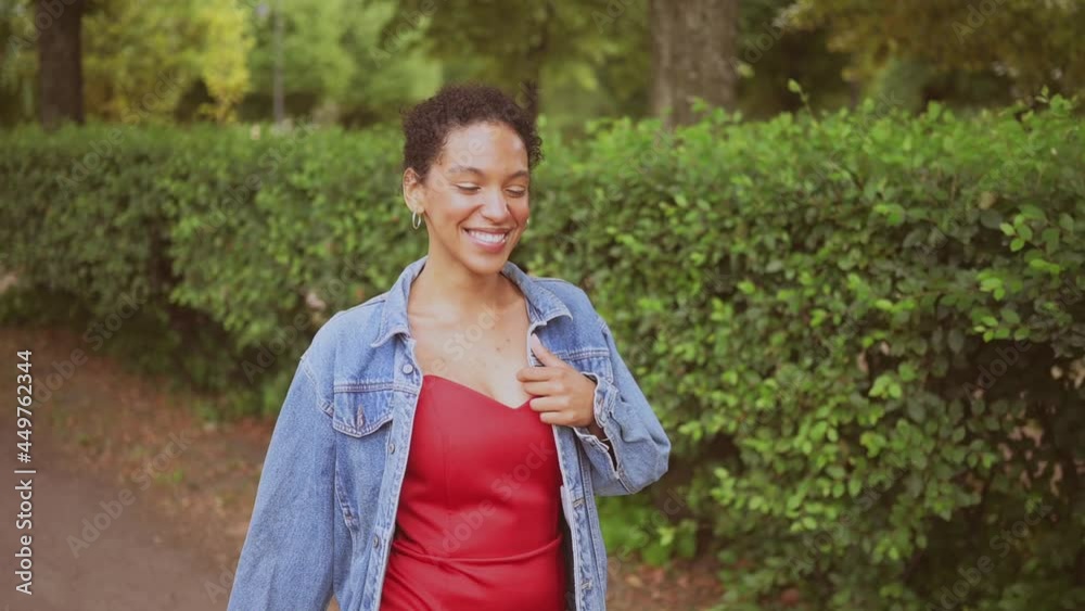 Fashion slow motion portrait of sensual attractive young naturally beautiful African American woman with black hair in jeans jacket and red dress walking in green nature parkland at summer Fashion slow motion portrait of sensual attractive young naturally beautiful African American woman with black hair in jeans jacket and red dress walking in green nature parkland at summer